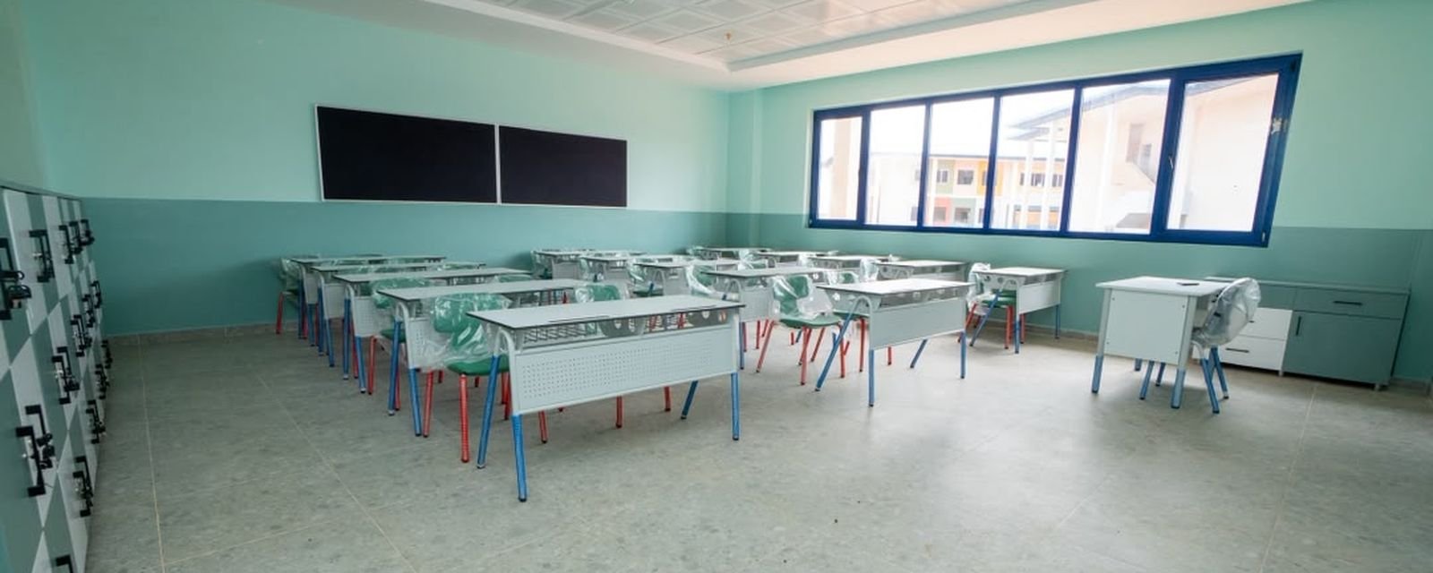 Une salle de classe meublée : tables doubles, tableaux et large baie vitrée